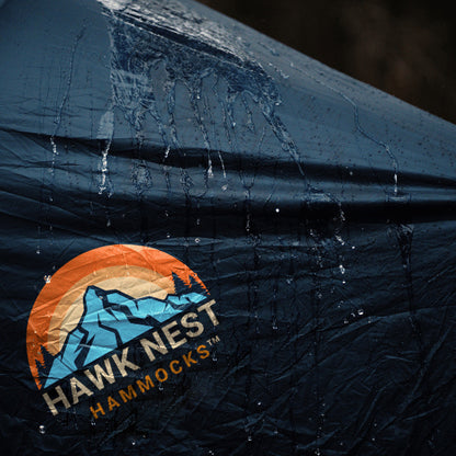 Ultralight hammock tent suspended between trees, ideal for outdoor camping and backpacking adventures. Compact, lightweight design ensures easy portability and setup. Perfect shelter solution for nature lovers seeking comfort and convenience in the wilderness. Water is shown beading off of the rain fly