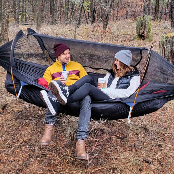 Ultralight hammock tent suspended between trees, ideal for outdoor camping and backpacking adventures. Compact, lightweight design ensures easy portability and setup. Perfect shelter solution for nature lovers seeking comfort and convenience in the wilderness. Two campers sitting in the hammock laughing